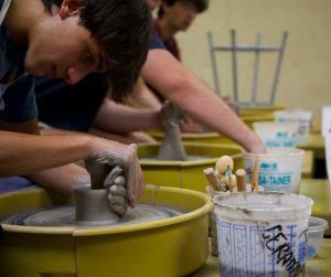 A young man is working at a pottery wheel in an art studio. He appears to be making a small cup. 