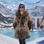 A woman with a fur coat, standing before a pool and mountains