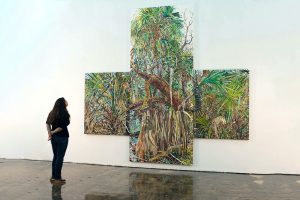 A woman stares at a painting of trees in a gallery.