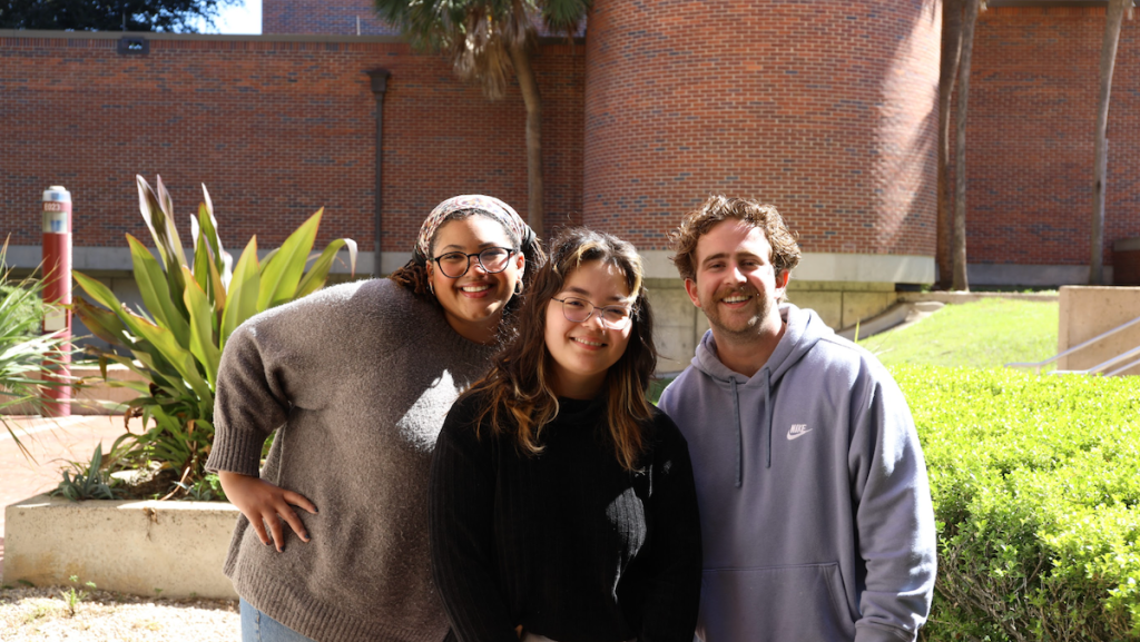 Three smiling art students pose for a photo in front of FSU's Fine Arts Building on a sunny day.