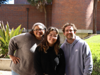 Three smiling art students pose for a photo in front of FSU's Fine Arts Building on a sunny day.