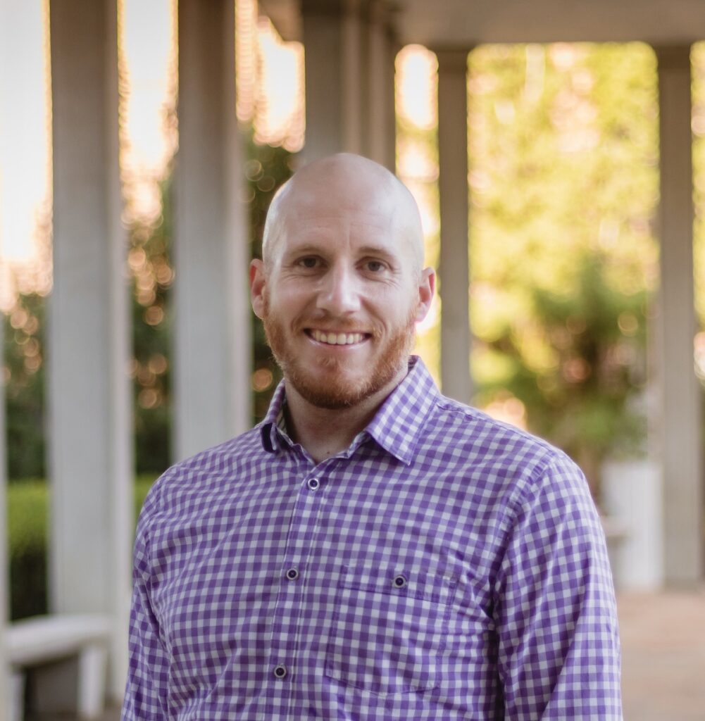 Jordan is wearing a plaid shirt smiling. He is standing outside in front of columns.