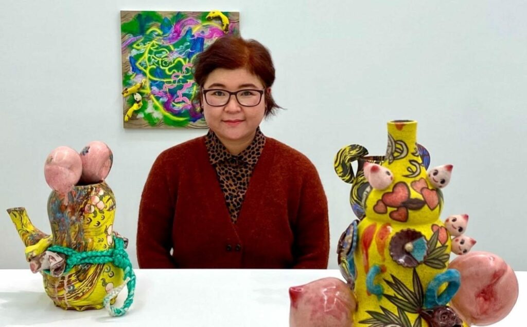 A woman in red poses in front of a colorful painting. In the foreground are two brightly colored sculptures.